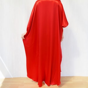 May include: A vibrant red, flowing caftan dress. The dress has a loose, oversized fit with wide sleeves and a floor-length hem. The fabric appears to be a smooth, silky material. The model is standing in front of a white wall and wooden floor.