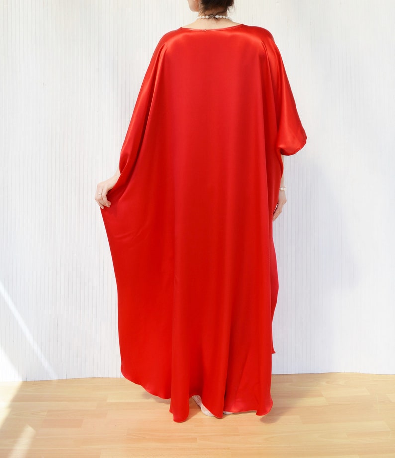 May include: A vibrant red, flowing caftan dress. The dress has a loose, oversized fit with wide sleeves and a floor-length hem. The fabric appears to be a smooth, silky material. The model is standing in front of a white wall and wooden floor.