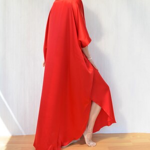 May include: A flowing, vibrant red dress with a wide, draped design. The dress features a loose, oversized fit with wide sleeves and a high-low hemline. The fabric appears to be a smooth, silky material. The model is wearing a pearl necklace and bracelet.