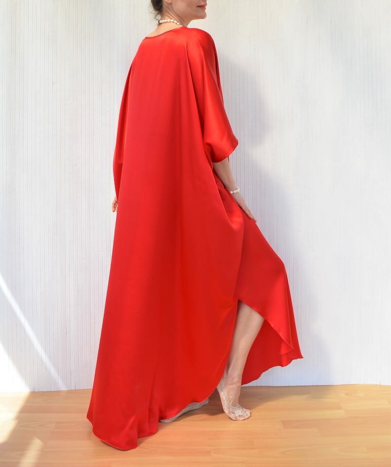 May include: A flowing, vibrant red dress with a wide, draped design. The dress features a loose, oversized fit with wide sleeves and a high-low hemline. The fabric appears to be a smooth, silky material. The model is wearing a pearl necklace and bracelet.