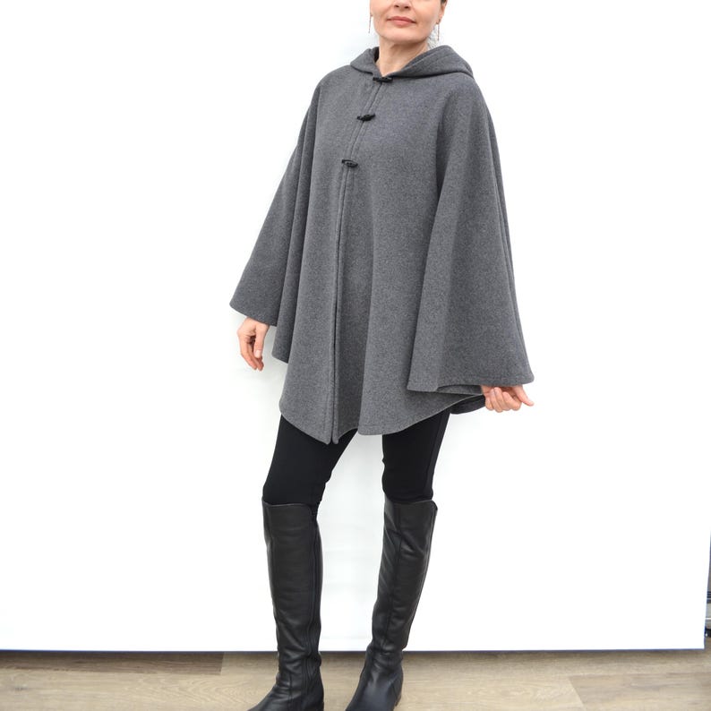 May include: A gray hooded cape with a long, flowing design. The cape has a zipper closure and two button closures at the neck. The model is wearing black leggings and black boots.