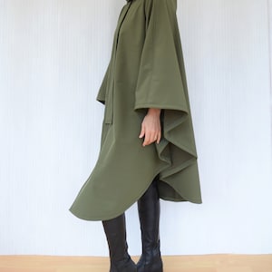 May include: Olive green hooded cape with a flowing design. The cape has a button closure and a hood. The model is wearing black boots. The cape is made of a soft, draping fabric.