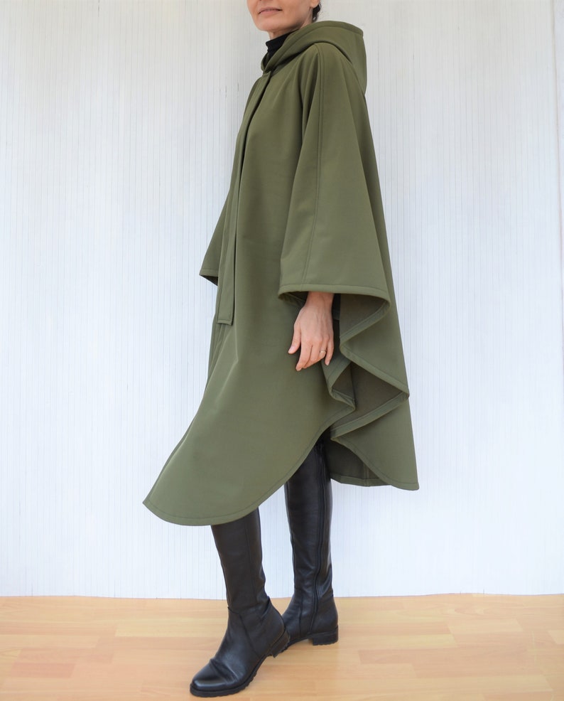 May include: Olive green hooded cape with a flowing design. The cape has a button closure and a hood. The model is wearing black boots. The cape is made of a soft, draping fabric.