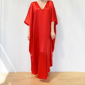 May include: A long, flowing red satin dress with a V-neck. The dress has wide sleeves and a loose, comfortable fit. The model is wearing a pearl necklace and bracelet. The background is a white wall and a wooden floor.