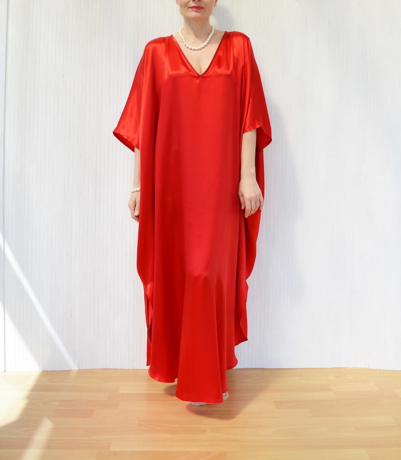 May include: A long, flowing red satin dress with a V-neck. The dress has wide sleeves and a loose, comfortable fit. The model is wearing a pearl necklace and bracelet. The background is a white wall and a wooden floor.