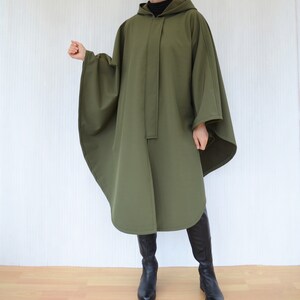 May include: An olive green hooded cape with a button closure down the front. The cape has wide sleeves and a rounded hem. The person is wearing black boots.