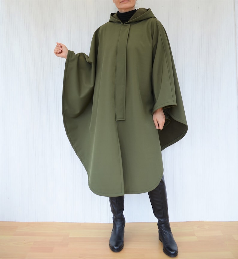May include: An olive green hooded cape with a button closure down the front. The cape has wide sleeves and a rounded hem. The person is wearing black boots.