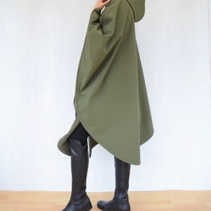May include: An olive green hooded poncho with a cape design. The knee-length poncho is worn with black leggings and tall black boots. The background is a plain white wall.