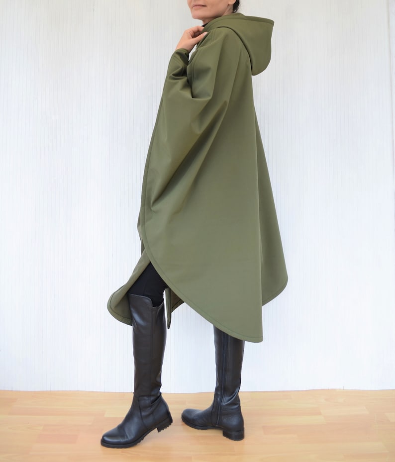 May include: An olive green hooded poncho with a cape design. The knee-length poncho is worn with black leggings and tall black boots. The background is a plain white wall.