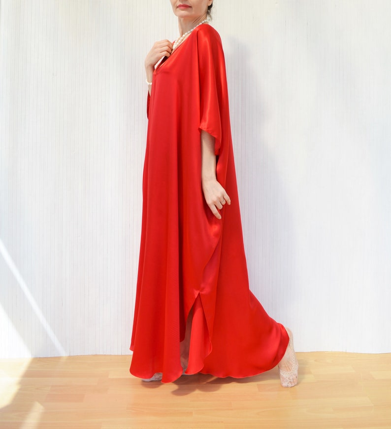 May include: A full-length, vibrant red caftan dress. The dress features a V-neckline and wide sleeves. The flowing fabric drapes to the floor, with a slit on one side. The dress is made of a shiny, silky material. The background is a white wall and a wooden floor.