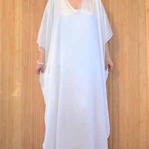 May include: A white, flowing kaftan dress with a V-neckline. The dress is made of a soft, silky fabric and has a relaxed fit.