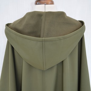 May include: Olive green hooded cape with a fleece-lined hood, displayed on a mannequin. The cape features a tailored design with a central seam and a flowing silhouette. The colour is a muted, earthy tone.