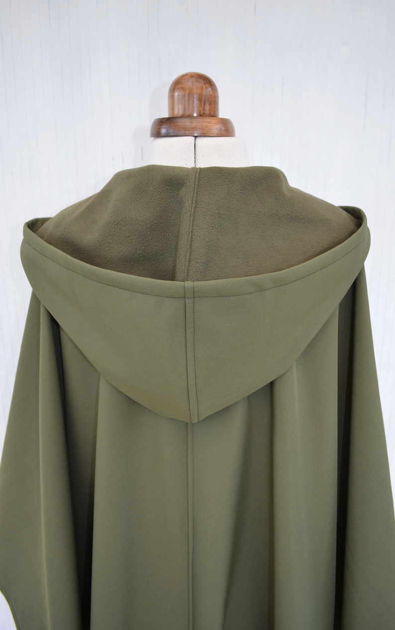 May include: Olive green hooded cape with a fleece-lined hood, displayed on a mannequin. The cape features a tailored design with a central seam and a flowing silhouette. The colour is a muted, earthy tone.