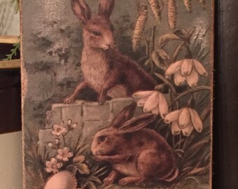 May include: Vintage Easter greeting card featuring two brown rabbits in a green grassy field with white flowers and a decorated Easter egg. The card has the text "Easter Greetings" at the bottom.