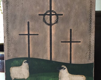 May include: A rustic wooden plaque featuring three black crosses, with a crown of thorns on the central cross. Two white sheep stand on a green hill in front of the crosses. The background is a neutral beige with a decorative border.
