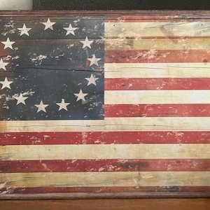 May include: Wooden framed wall art featuring the American flag with 13 stars in a circle and red and white stripes. The flag has a distressed, vintage appearance, with visible wood grain and a dark blue field.