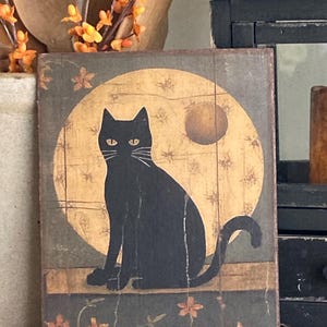 May include: A black cat sits in front of a full moon with a smaller moon in the background. The cat is silhouetted against the moon. The background is a dark green with orange flowers.