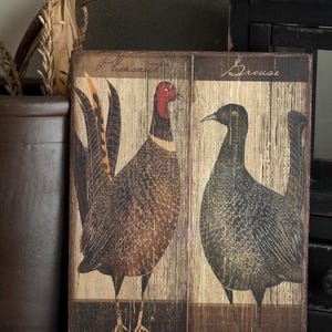 May include: Wooden wall art featuring two birds, a pheasant and a grouse, with the words "Pheasant" and "Grouse" above each bird. The birds are depicted in a rustic, vintage style with brown, red, and black tones. The background has a wood grain texture.