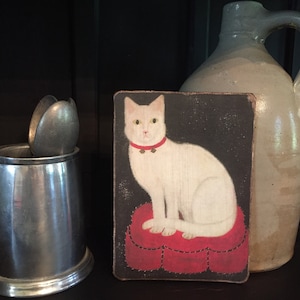 May include: A white cat with a red collar and bells sits on a red heart-shaped cushion. The cat is painted on a dark background and framed in a wooden frame.