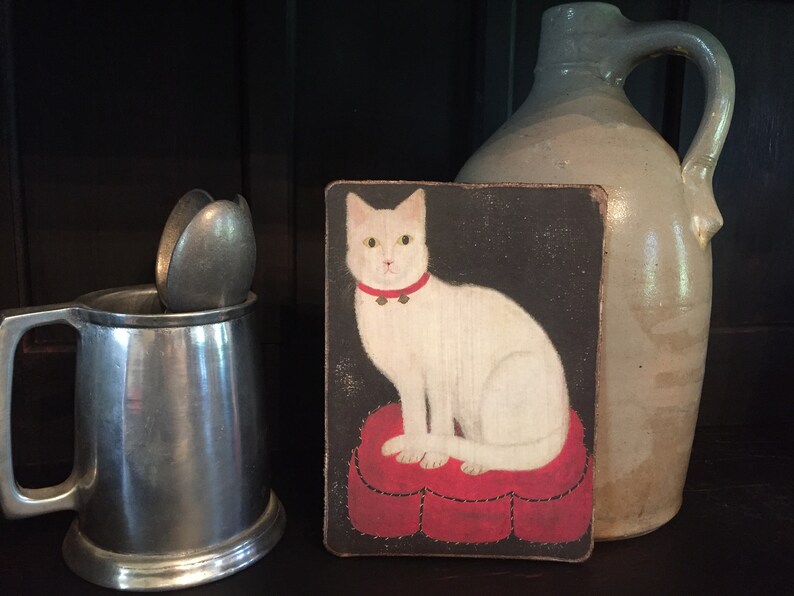 May include: A white cat with a red collar and bells sits on a red heart-shaped cushion. The cat is painted on a dark background and framed in a wooden frame.