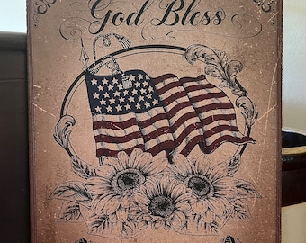 Könnte beinhalten: Ein Schild im Vintage-Stil mit dem Text "God Bless America" in einer dekorativen Schriftart. Das Schild zeigt eine stilisierte amerikanische Flagge mit Sonnenblumen und einem dekorativen Rand. Das Schild misst 20,32 x 25,4 cm.