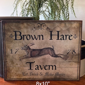 May include: A rectangular wooden sign with the words "Brown Hare Tavern" and a running hare illustration. The sign is 8x10 inches and features the text "Eat, Drink & Make Merry". The sign has a vintage, distressed look.