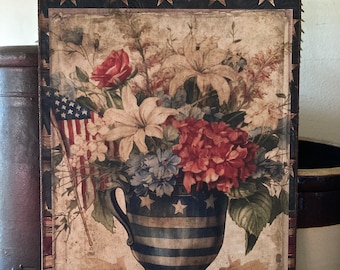 Puede incluir: Un letrero de madera de estilo vintage con un tema patriótico. Presenta un ramo de flores rojas, blancas y azules en un jarrón a rayas azul y blanco con estrellas. El jarrón está sobre una superficie de madera con un fondo a rayas rojo, blanco y azul. El letrero también presenta una pequeña bandera estadounidense y la palabra "America" en la parte inferior.