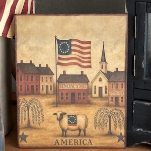 May include: A rustic painting depicts an American flag and a colonial village scene. The artwork features a sheep with a flag design, a general store, a church, and the word "AMERICA" at the bottom. The color palette includes muted reds, browns, and creams.