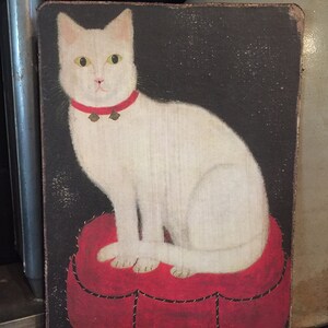 May include: A white cat with a red collar and bells sits on a red heart-shaped cushion against a dark background.