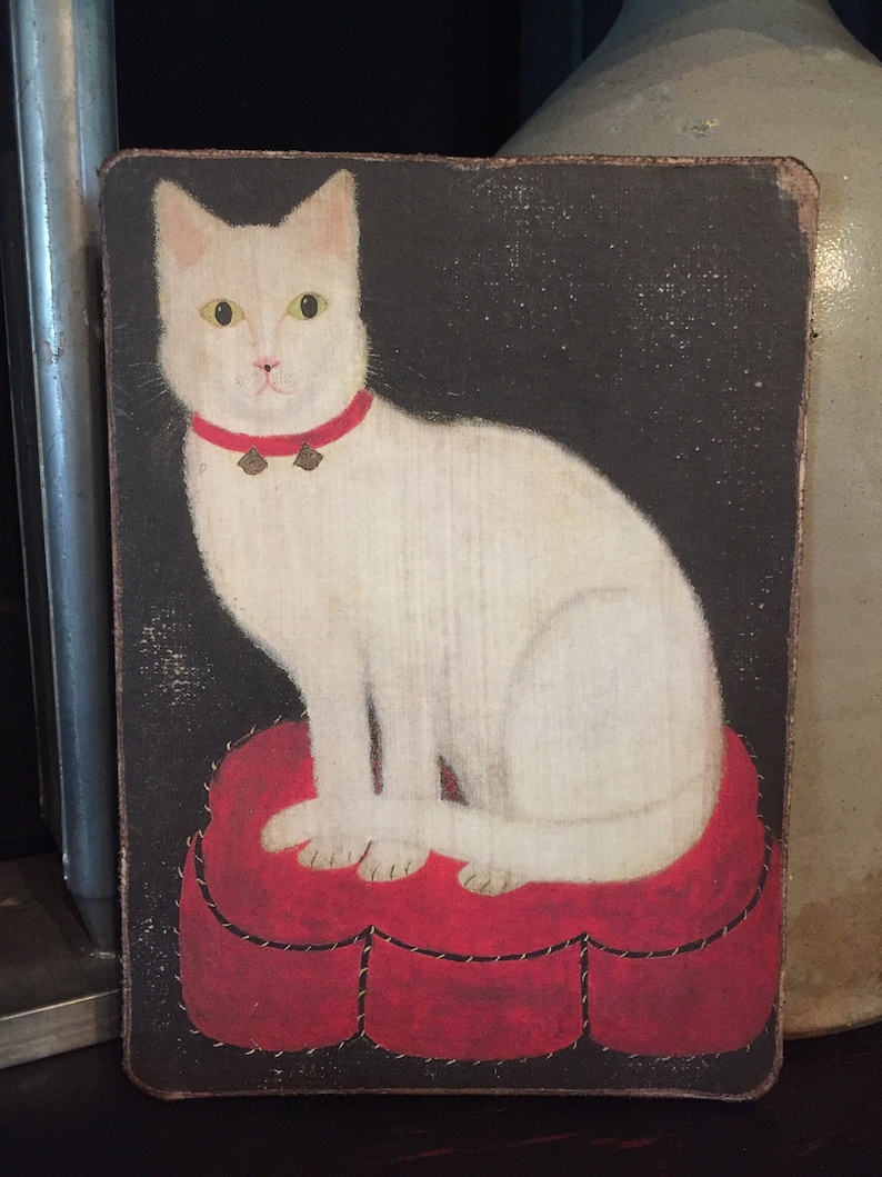 May include: A white cat with a red collar and bells sits on a red heart-shaped cushion against a dark background.