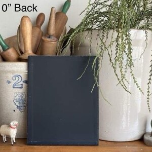 May include: A black 8x10 inch chalkboard with a white frame, sitting on a wooden surface. There are two small white sheep figurines in the foreground. The chalkboard is in front of a white ceramic crock with a blue design and a white ceramic crock with a green plant.