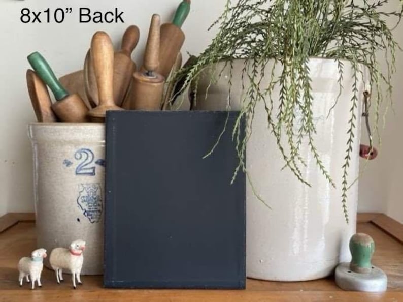 May include: A black 8x10 inch chalkboard with a white frame, sitting on a wooden surface. There are two small white sheep figurines in the foreground. The chalkboard is in front of a white ceramic crock with a blue design and a white ceramic crock with a green plant.