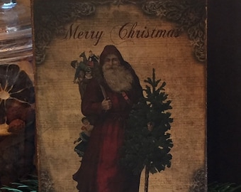 May include: A rectangular Christmas sign featuring an illustration of Santa Claus in a red robe carrying a Christmas tree and a sack. The sign has the words "Merry Christmas" in elegant script and is framed with decorative flourishes.