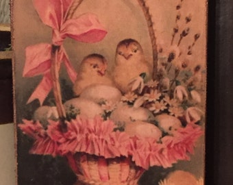May include: Vintage Easter-themed wooden sign featuring a basket filled with eggs, chicks, and flowers. A large pink bow adorns the basket's handle. The words "Easter Greetings" are written in red script at the top. The sign has a distressed, aged appearance.