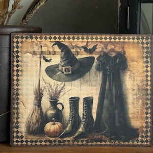 May include: A framed Halloween-themed print featuring a witch's hat, dress, boots, and broom. The artwork has a vintage, rustic style with a muted color palette of black, brown, and orange. A small pumpkin and vase of flowers are also depicted.