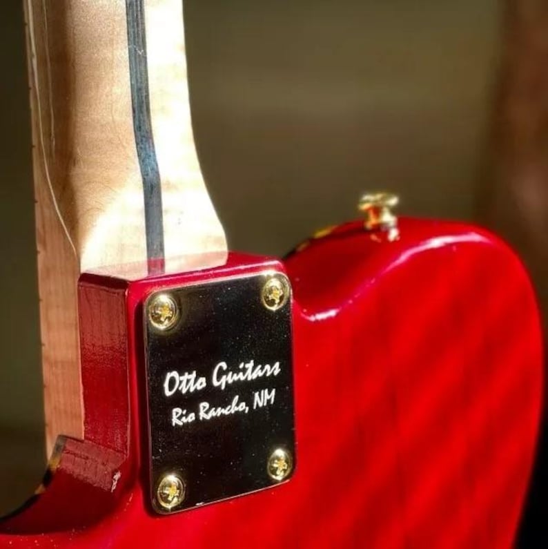 May include: A close-up of a red electric guitar body with a wooden neck. A black plate with gold screws is attached to the body, displaying the text "Otto Guitars Rio Rancho, NM". The guitar has a glossy finish.