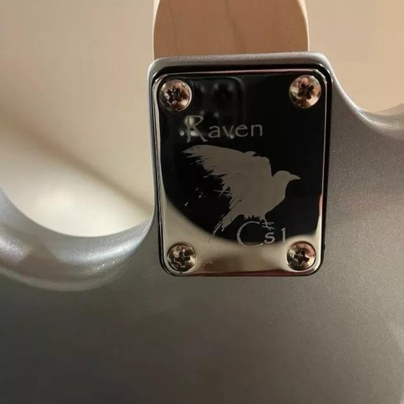 May include: Close-up of a silver guitar neck plate with the word "Raven" and a bird silhouette etched on it. The plate is secured with four gold screws. The guitar body is a metallic silver colour.