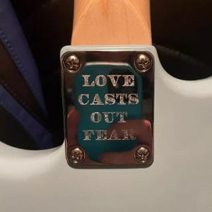 May include: A close-up shot of a silver metal plate with the words "LOVE CASTS OUT FEAR" engraved on it. The plate is secured with screws to a white surface. The background is blurred.