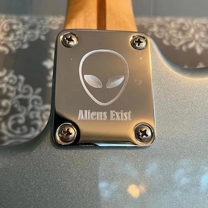 May include: A close-up of a guitar neck plate with the text "Aliens Exist" below an alien head graphic. The plate is silver and secured with four screws. The guitar body is a light blue colour.