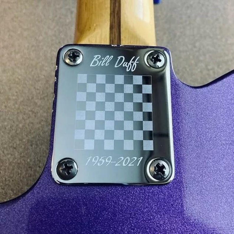 May include: Detail of a purple electric guitar's neck joint. A silver plate is secured with screws, displaying the engraved text "Bill Duff", a checkered design, and the dates "1959-2021".