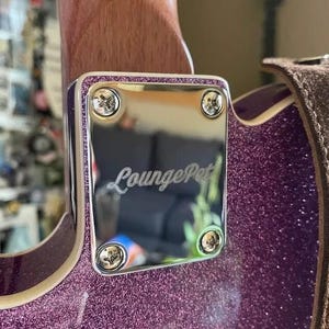 May include: Detail of a guitar's neck joint, showing a polished silver plate with the cursive text "LoungePet". The plate is fixed with four screws and framed by a purple glitter border. The guitar's body has a matching purple glitter finish.