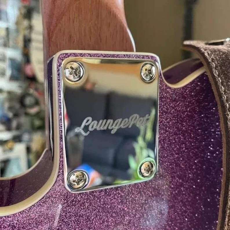May include: Detail of a guitar's neck joint, showing a polished silver plate with the cursive text "LoungePet". The plate is fixed with four screws and framed by a purple glitter border. The guitar's body has a matching purple glitter finish.