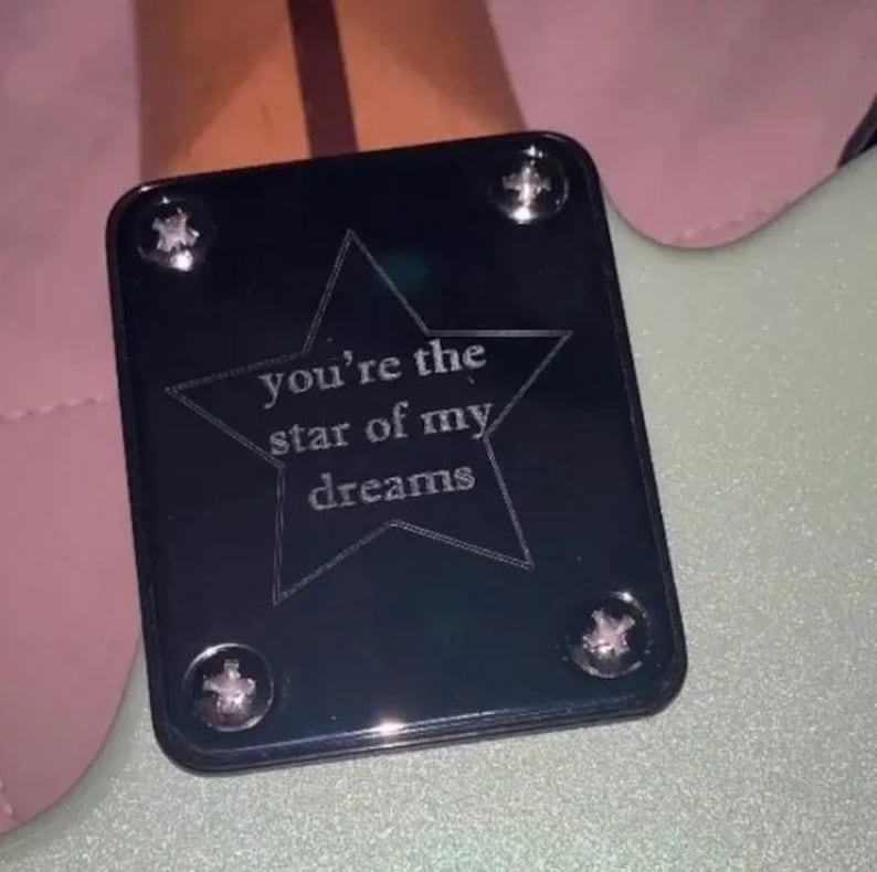 May include: A black guitar neck plate with the words "you're the star of my dreams" inside a star outline. It is secured with four silver screws. The background is light green and pink.