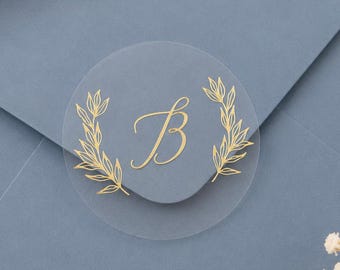 May include: A circular transparent sticker featuring a gold foil letter B in a cursive script, flanked by two delicate gold foil botanical leaf sprigs, centered on a dusty blue envelope flap with a smooth matte finish and clean edges.