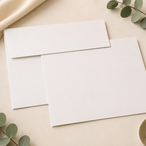 May include: A plain white rectangular envelope and a matching white rectangular card. The envelope is positioned horizontally with the flap folded down, and the card is placed partially underneath the envelope, showing a clean, smooth surface.