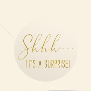 May include: A clear round sticker with gold text that says "Shhh... It's a surprise!"