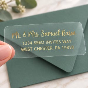 May include: A rectangular clear adhesive label featuring gold foil text that reads Mr. & Mrs. Samuel Baron, 1234 Seed Invites Way, West Chester, PA 19810. The label has rounded corners and is designed for use as a return address sticker on envelopes.