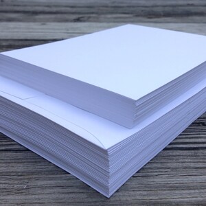 May include: A stack of crisp white rectangular envelopes arranged in two tiers. The bottom layer consists of a larger pile of envelopes, while a smaller, thinner stack of envelopes is placed directly on top, showcasing clean edges and smooth surfaces.