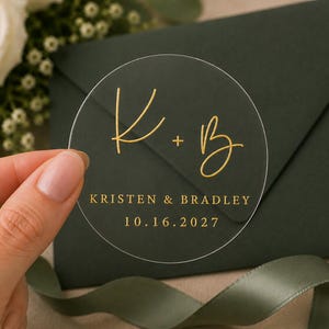 May include: A circular transparent sticker featuring gold lettering with the initials K + B, the names KRISTEN & BRADLEY, and the date 10.16.2027. The design uses a combination of a script font for the initials and a serif font for the text.