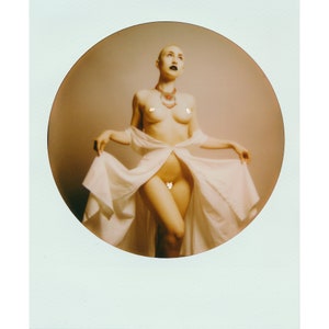 Könnte beinhalten: Eine Frau mit rasiertem Kopf und dunklem Make-up trägt ein weißes Gewand und eine rot-goldene Halskette. Sie steht mit ausgestreckten Armen und blickt nach oben.